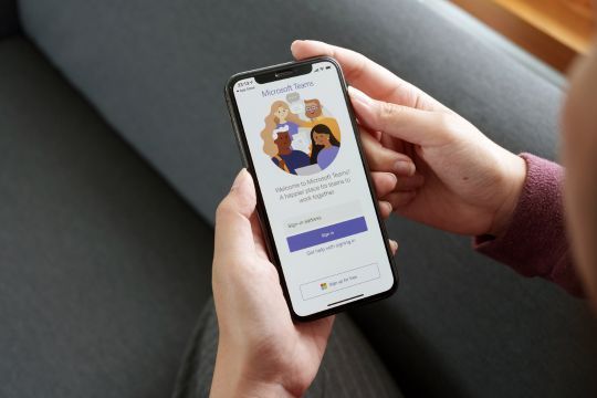Eine Person hält ein Smartphone in der Hand, auf dem der Anmeldebildschirm der Microsoft Teams App angezeigt wird. Der Text auf dem Bildschirm bietet Optionen für das Anmelden oder die Erstellung eines Kontos. Der Hintergrund besteht aus einem grauen Sofa.