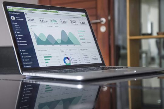 Ein Laptop, der ein Business-Dashboard mit verschiedenen Grafiken und Diagrammen anzeigt, steht auf einem reflektierenden Tisch. Im Hintergrund ist ein modernes Büro mit einem Holzrahmen und einer Glaswand zu sehen.