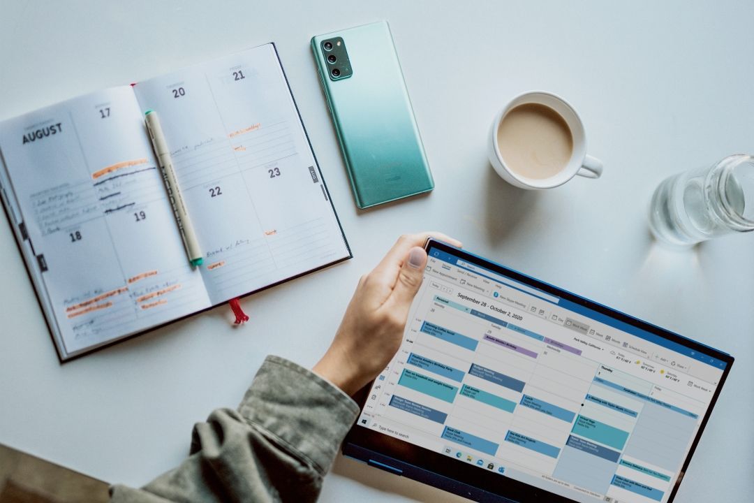 Person hat ein Tablet in der Hand, daneben liegen ein Kalender, ein handy und eine Kaffeetasse